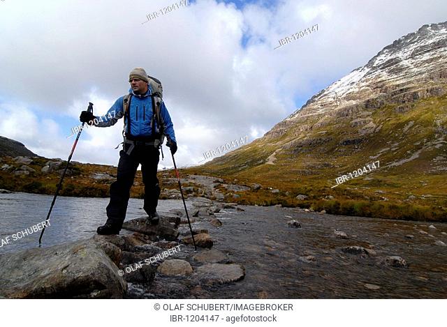Hiker with trekking poles and backpack crossing a river in the Scottish mountains, Scottish Highlands, Stuca `Choir Dhuibh Beig, Liathach, Torridon, Scotland