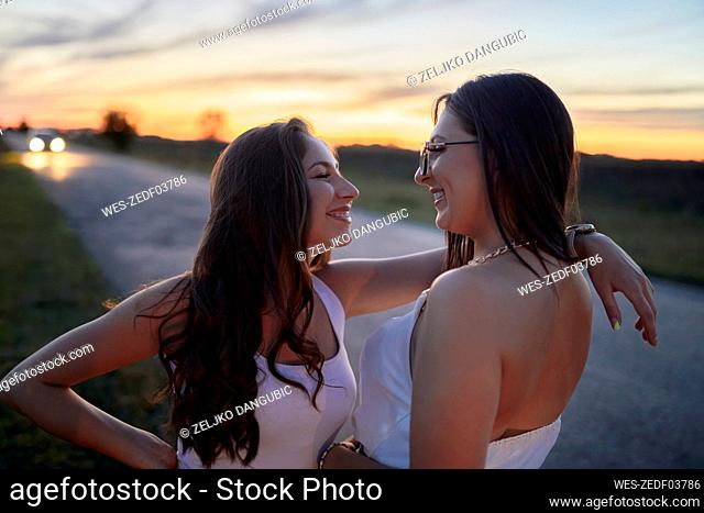 Smiling female friends looking at each other while standing on road against sky at sunset