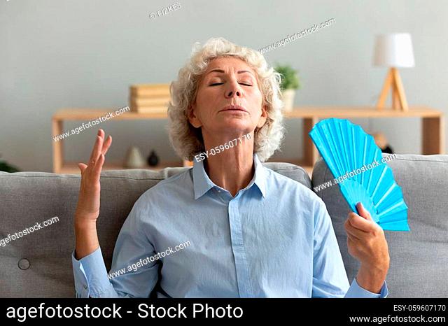 Overheated mature woman sit on couch in living room breathe fresh air from hand waver, thirsty sick elderly female relax on sofa at home wave using fan suffer...