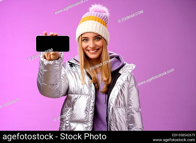 Confident friendly good-looking blond girl in outdoor silver glittering jacket hat hold smartphone horizontal showing mobile phone display assertive smile...