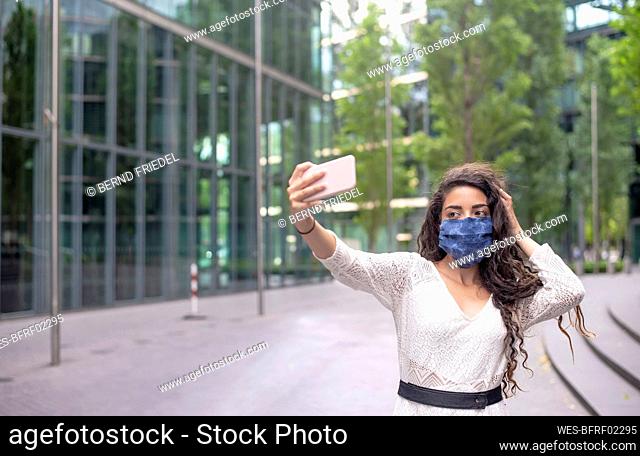 Young woman wearing mask taking selfie with smart phone while standing on footpath in city