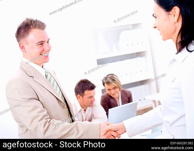 Businessman and businesswoman shaking hands in office, colleagues working on laptop in background