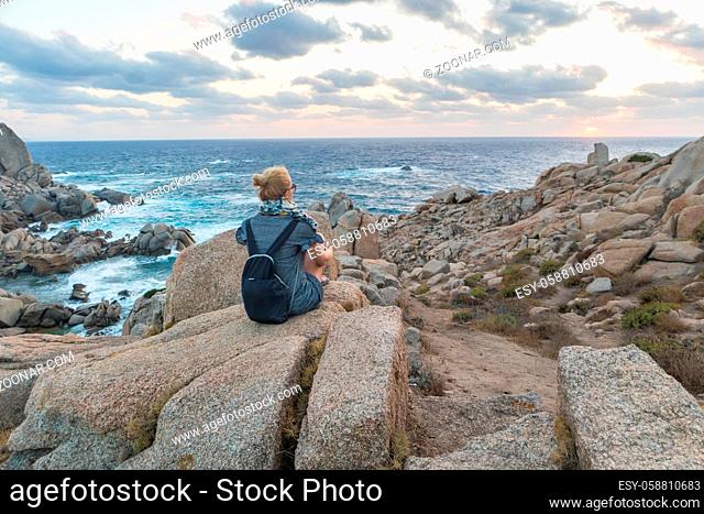 Solitary young female traveler watches a beautiful sunset on spectacular rocks of Capo Testa, Sardinia, Italy, a popular summer traveling destination in Europe