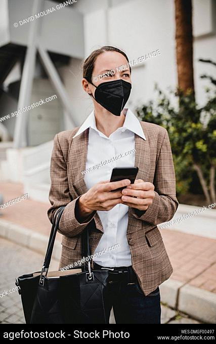 Businesswoman wearing protective face mask holding smart phone standing against building while looking away