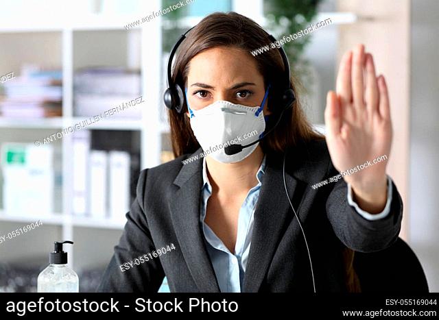 Serious telemarketer woman gesturing stop avoiding covid-19 with mask looking at camera sitting at the office