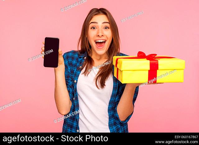 Wow, online delivery order. Surprised pretty girl in checkered shirt holding gift box, cell phone and looking with amazement