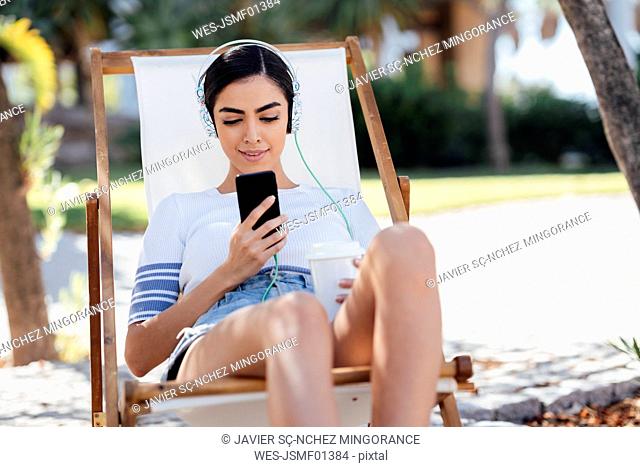 Young woman sitting in a deckchair wearing headphones and using smartphone