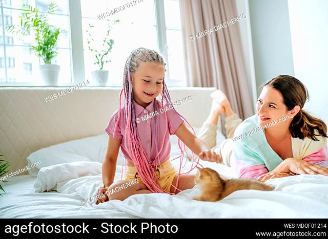 Mother looking at daughter playing with ginger kitten on bed