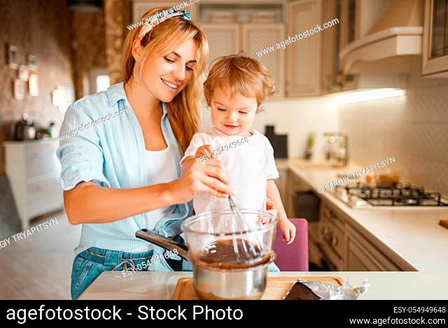 Young mother with her daughter mixing melted chocolate in a bowl. Cute woman and little girl cooking on the kitchen. Happy family prepares sweet dessert at the...