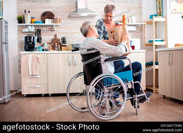 Senior woman taking grocery paper bag from handicapped husband in wheelchair. Mature people with fresh vegetables from market