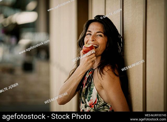 Happy mature woman eating apple by wall