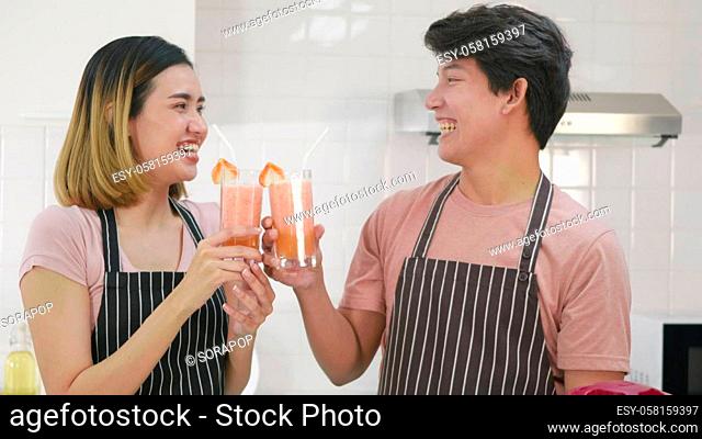 Happy Asian beautiful young family couple husband and wife enjoying drinking fresh apple smoothie juice in kitchen on glass together in morning at home