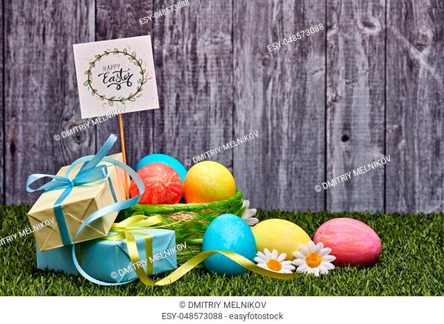 Colored easter eggs, gift boxes and flowers lying on a green grass on a grey wooden background with copy space. Greeting card, Easter background