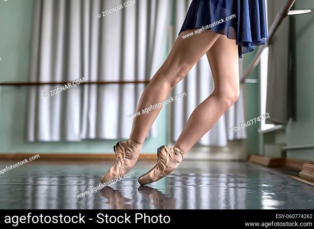 Legs of a ballet dancer in a blue dance wear who stands on pointes next to the ballet barre. She wears beige pointe shoes