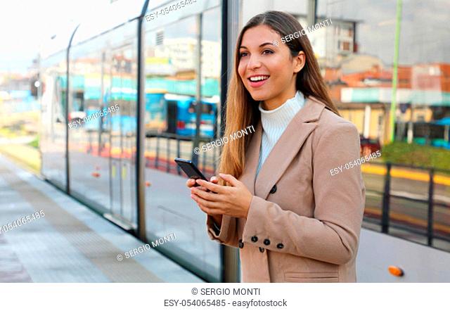 City transport. Happy beautiful woman holding cellular at tram stop. Smiling business woman satisfied with online ticket service paying for electric transport...