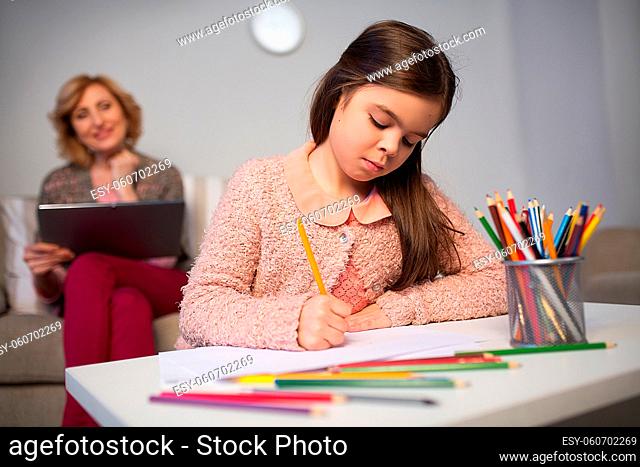 Little girl drawing house with color pencils. Graany sitting behind on sofa