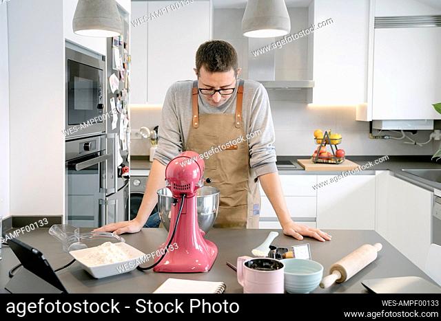 Mid adult man with apron looking at stand mixer while preparing food at home