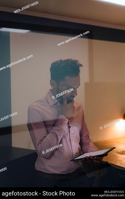 Thoughtful businessman using tablet PC seen through glass in office