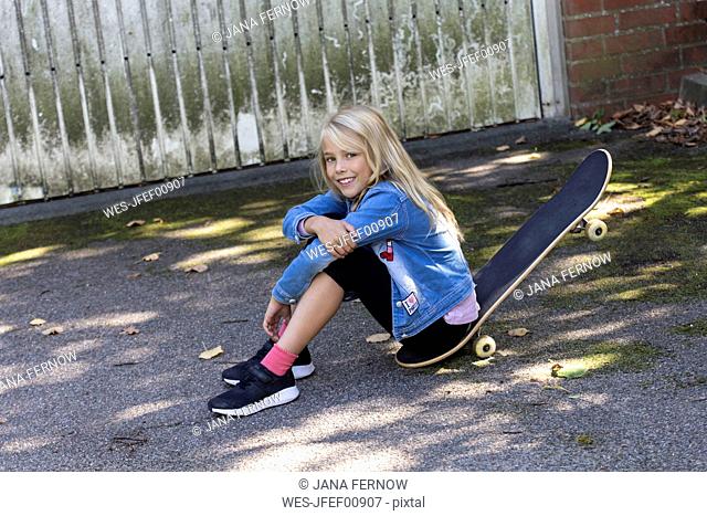 Portrait of smiling blond girl sitting on her skateboard outdoors