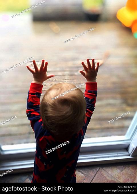 A toddler, probably attracted by the objects outside, looking out on a wooden deck through a giant glass door, Nova Scotia, Canada