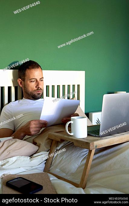 Man with laptop analyzing document while lying on bed