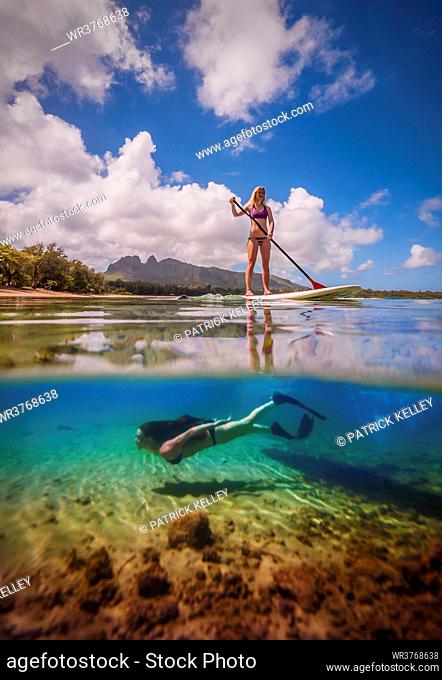 An athletic woman rides a stand-up paddle-board as another woman free-dives below her, Hawaii, United States of America, Pacific