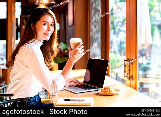 Quarantine Young asian woman wearing smart casual clothes work at home in living room using laptop and drinking hot coffee and croissant while city lockdown for...