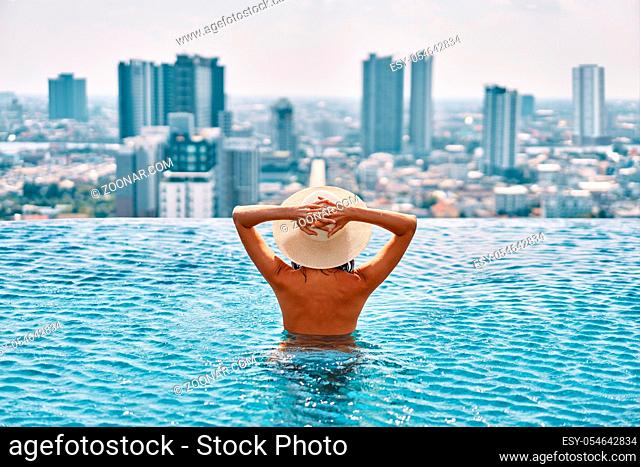 Back view of young woman in hat relaxing in swimming pool on the roof top of hotel and enjoy cityscape. Summer vacation, luxury concept