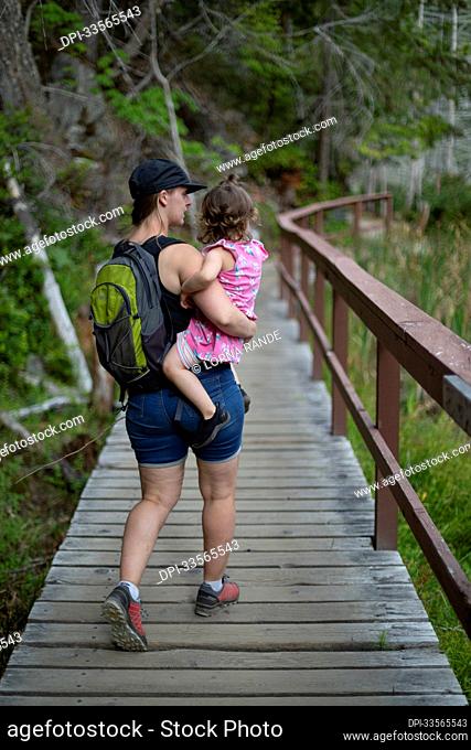 Mother carries her young daughter as they walk down a trail on a hike together in Smuggler Cove Marine Provincial Park along the Sunshine Coast of BC