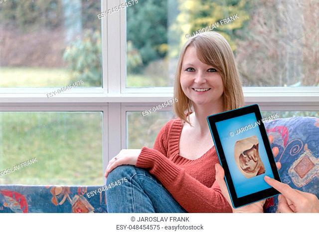 Smiling young woman is sitting at home and another female hands are showing space shot of the unborn child in the tablet screen