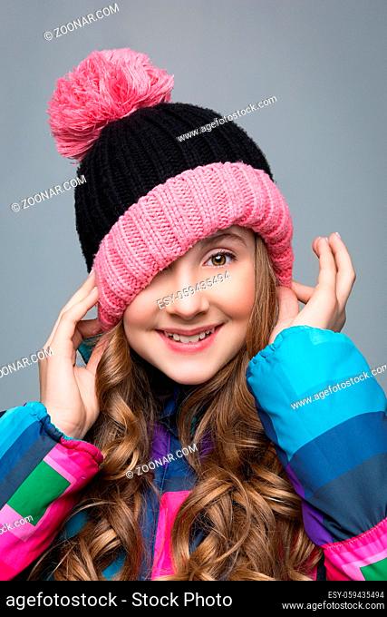 Beautiful happy teen girl with long curly hair in pink wool hat and bright warm coat. Studio shot over grey background. Copy space