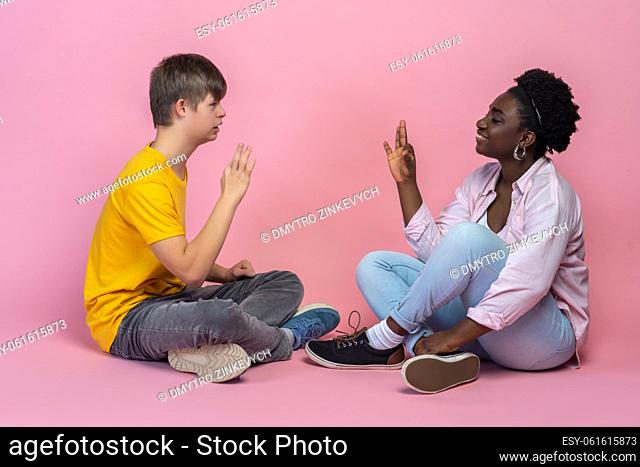 Sign language. Guy with down syndrome and dark-skinned woman sideways to camera sitting on floor opposite each other showing signs