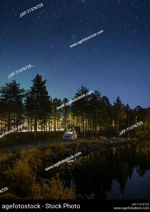 Camping with VW Bulli Campervan on the lake shore with trees under the starry sky at night in Lapland, Sweden
