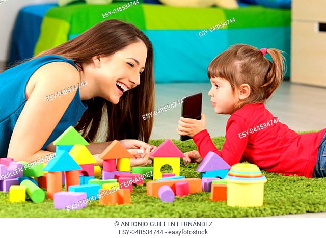 Toddler showing a smart phone to her mother lying on a carpet in a room