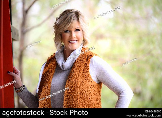 A 60 year old blond woman smiling at the camera in an outdoor setting