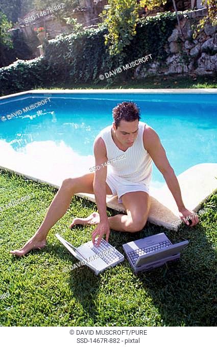 Young man sitting beside a swimming pool using a laptop
