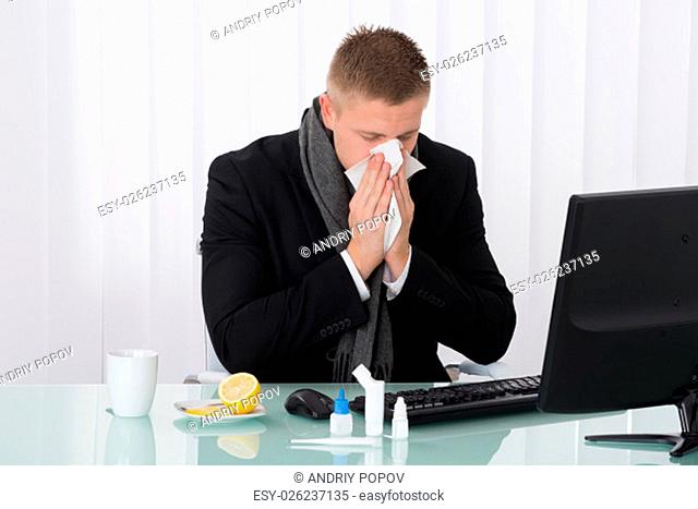 Sick Businessman Blowing His Nose In Office With Medicines On Desk