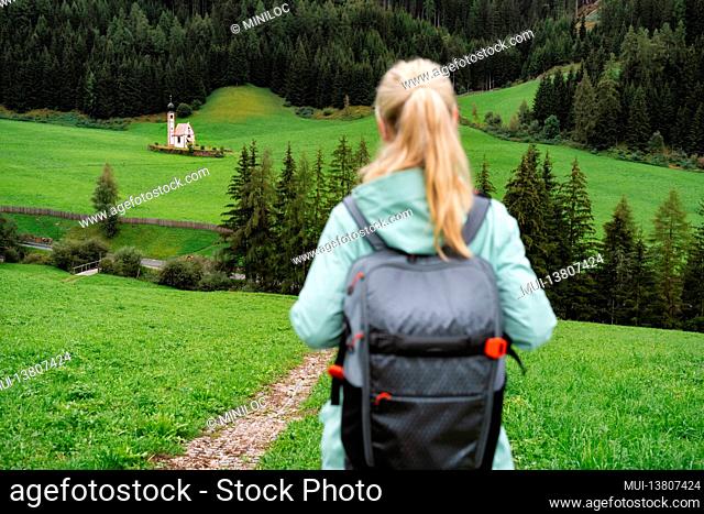 Hiking woman with backpack enjoy the view of St. Johann church in Val di Funes valley, Dolomites, Italy, Europe