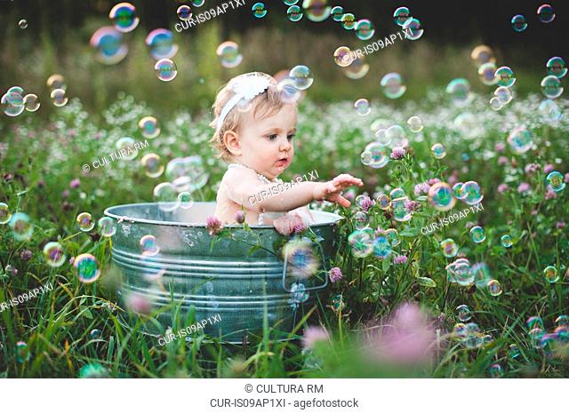 Baby girl in tin bathtub in meadow reaching for floating bubbles