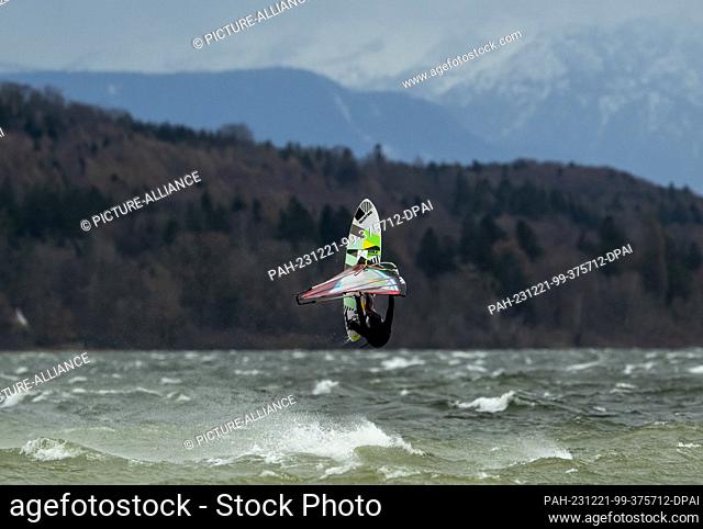 21 December 2023, Bavaria, Munich: A surfer takes advantage of the strong wind on Lake Ammersee near Herrsching to jump with his board against the backdrop of...