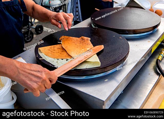 A close-up view on the hands of a street food seller using a wooden spatula to fold freshly prepared crepes on a griddle outdoors