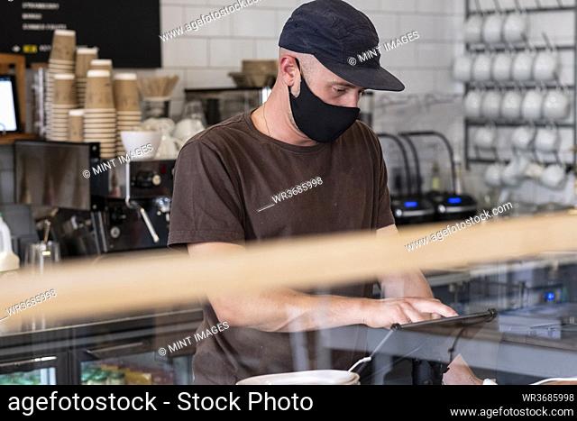 Male barista wearing black baseball cap and face mask working behind counter