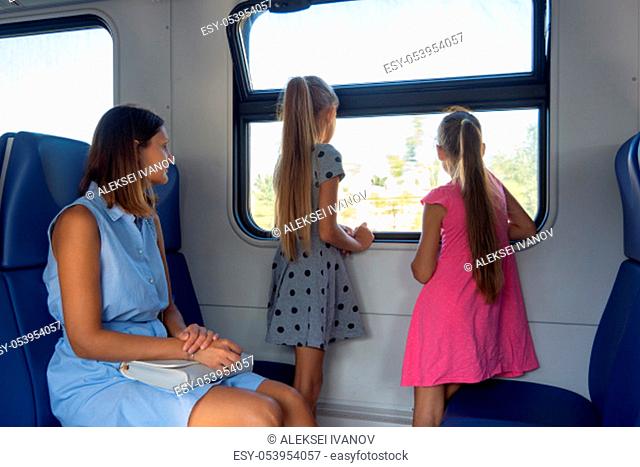 Mom and two daughters ride in an electric train, children standing looking out the window