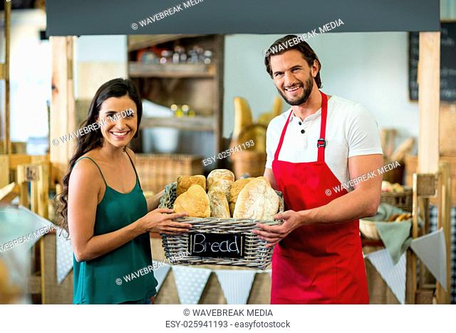 Portrait of smiling male staff and woman holding a basket of baguettes at counter