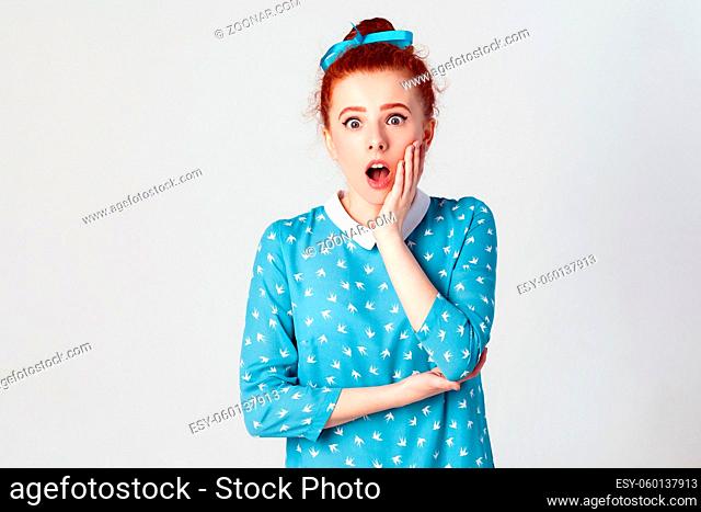 Ginger young girl screaming with shock, holding hands on her cheeks. Isolated studio shot on gray background