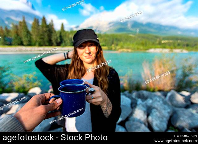 Selective focus of two persons clinching their cups of coffee and tea together while standing in front of a nature scenery with a river