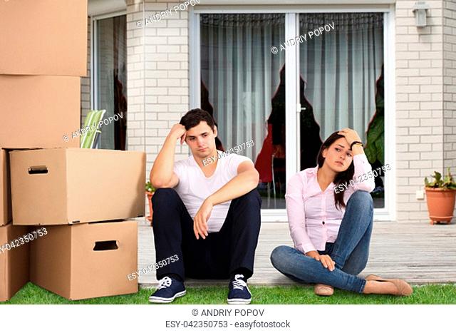 Upset Couple Sitting Near Stacked Cardboard Boxes In Front Of Their House
