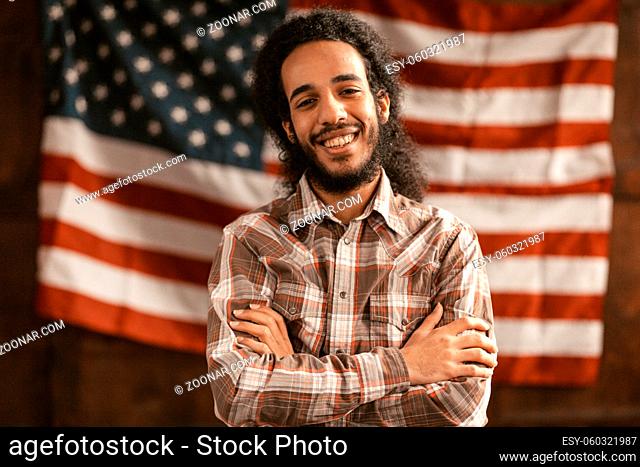 The dark-skinned person Arab exterior cheerfully laughs with his arms crossed. Patriotic American man rejoices in success on American flag background