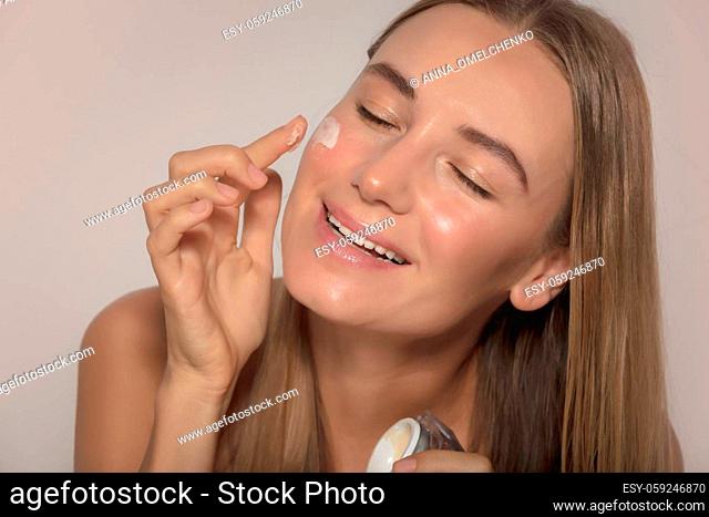 Portrait of a Nice Female with Closed Eyes of Pleasure Applying Moisturizer Cream on Face. Natural Cosmetics. Isolated on Beige Background