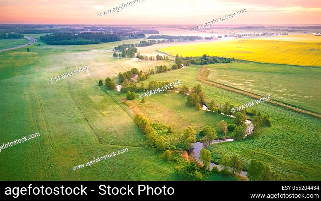 Spring morning aerial rural panorama. Sunrise over green and yellow blooming colza fields. Clouds of fog and small river with trees on riverbank